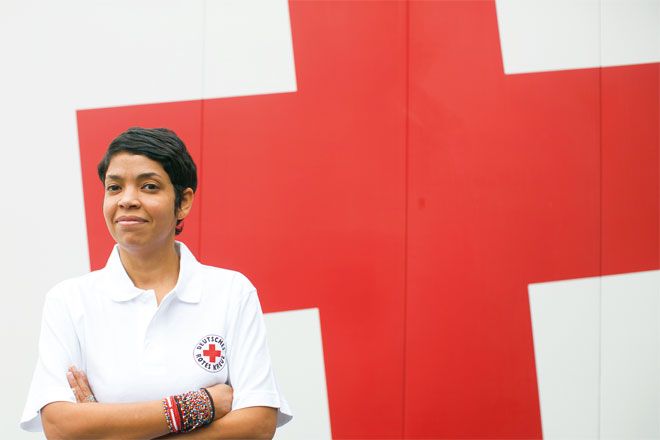 Bild: Eine junge DRK-Helferin steht vor einer weißen Wand mit einem großen, roten Kreuz. Sie blickt direkt in die Kamera und wirkt insgesamt zuversichtlich und gelassen.