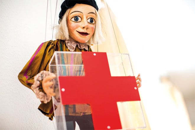 Bild: Eine Marionette hält vor sich ein großes, rotes Kreuz in den Händen.